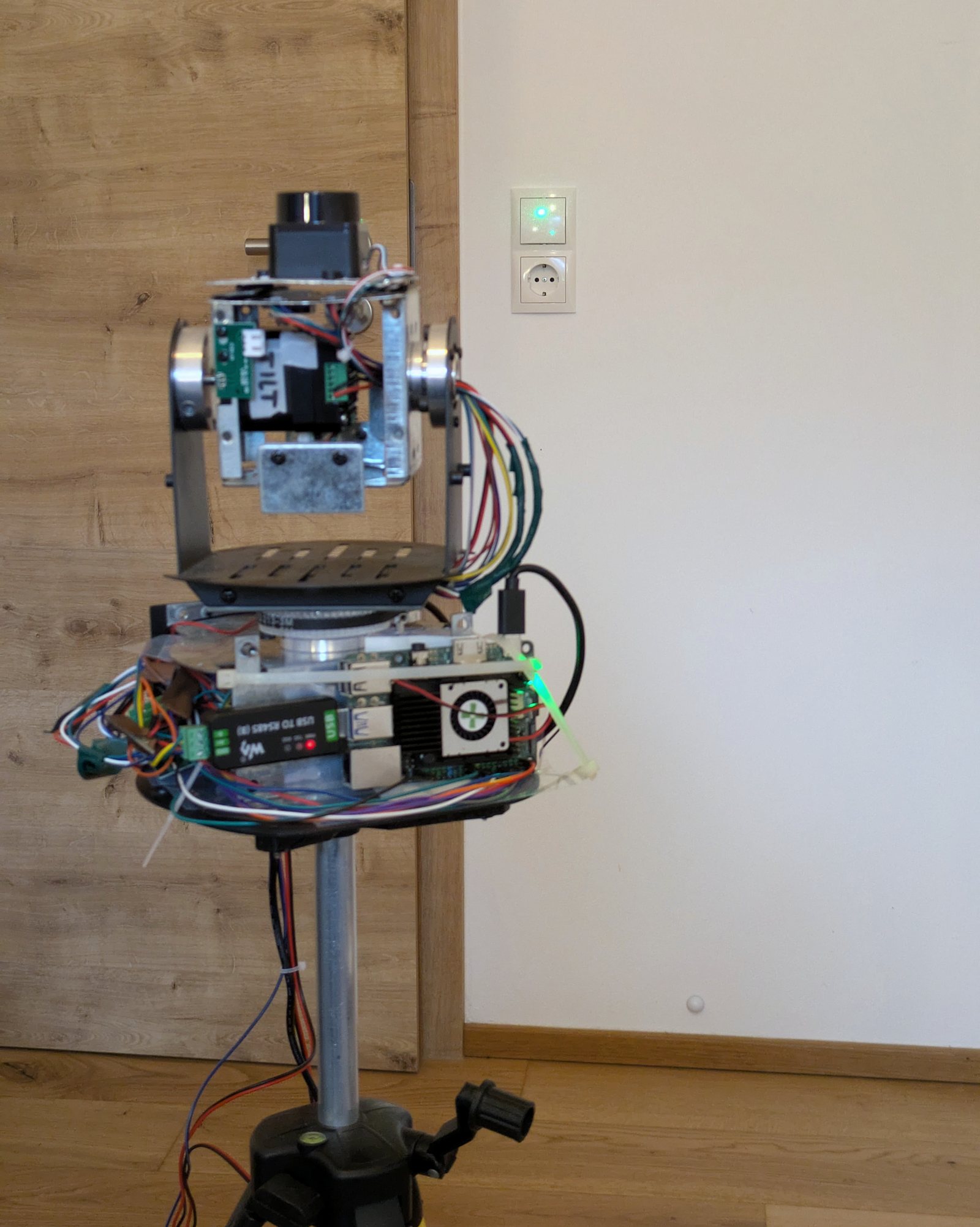 Funktionsprototyp von Aurintex Beam auf einem Stativ mit offener Elektronik. Ein grüner Laserpunkt liegt auf dem Lichtschalter neben einer Holztür.